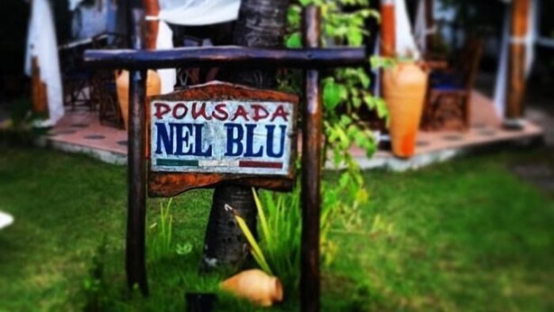 A sign with the text "Pousada Nel Blu" in front of a building with a red-tiled roof and white curtains. - Pousada Nel Blu (Itaparica)