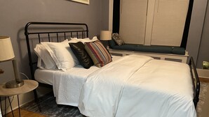 A neatly made bed with white bedding, a bedside lamp, and a window with blinds. - Beaumont Nurses or Dr’s need quarantine site?<br>Downtown Royal Oak, charming 2bd (Royal oak)