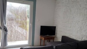 Een woonkamer met een donkergrijze bank, een flatscreen televisie op een houten dressoir, en een groot raam met gordijnen.