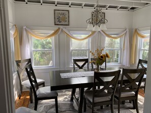Une salle à manger avec une table en bois foncé, quatre chaises et un lustre. De grandes fenêtres sont ornées de rideaux, et un tableau encadré est accroché au mur.
