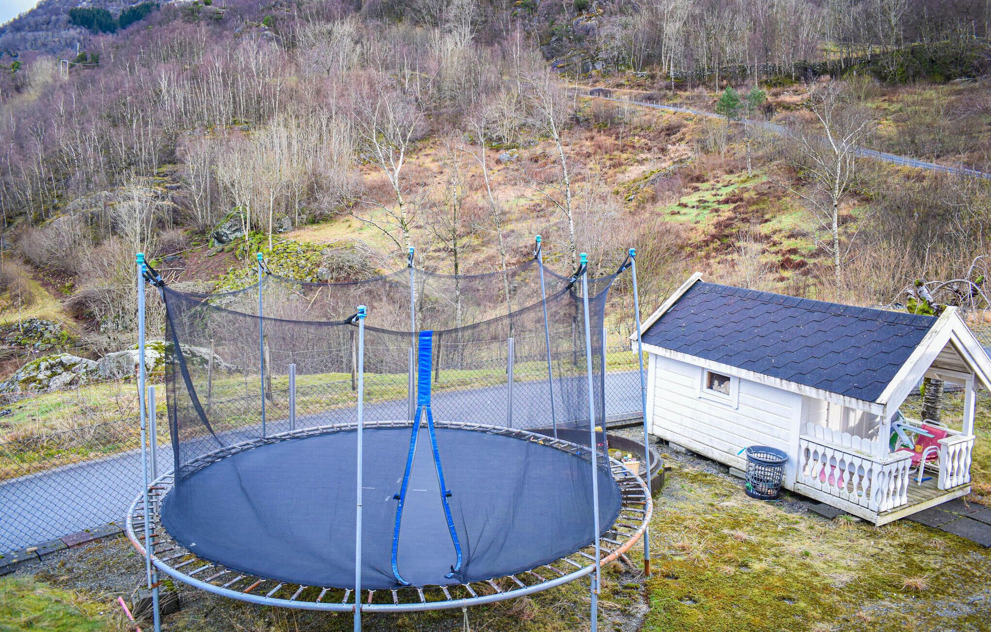 Ein Trampolin, umringt von einem Sicherheitsnetz, eine kleine weiße Holzhütte mit dunklem Dach und ein bewaldeter Hang im Hintergrund.