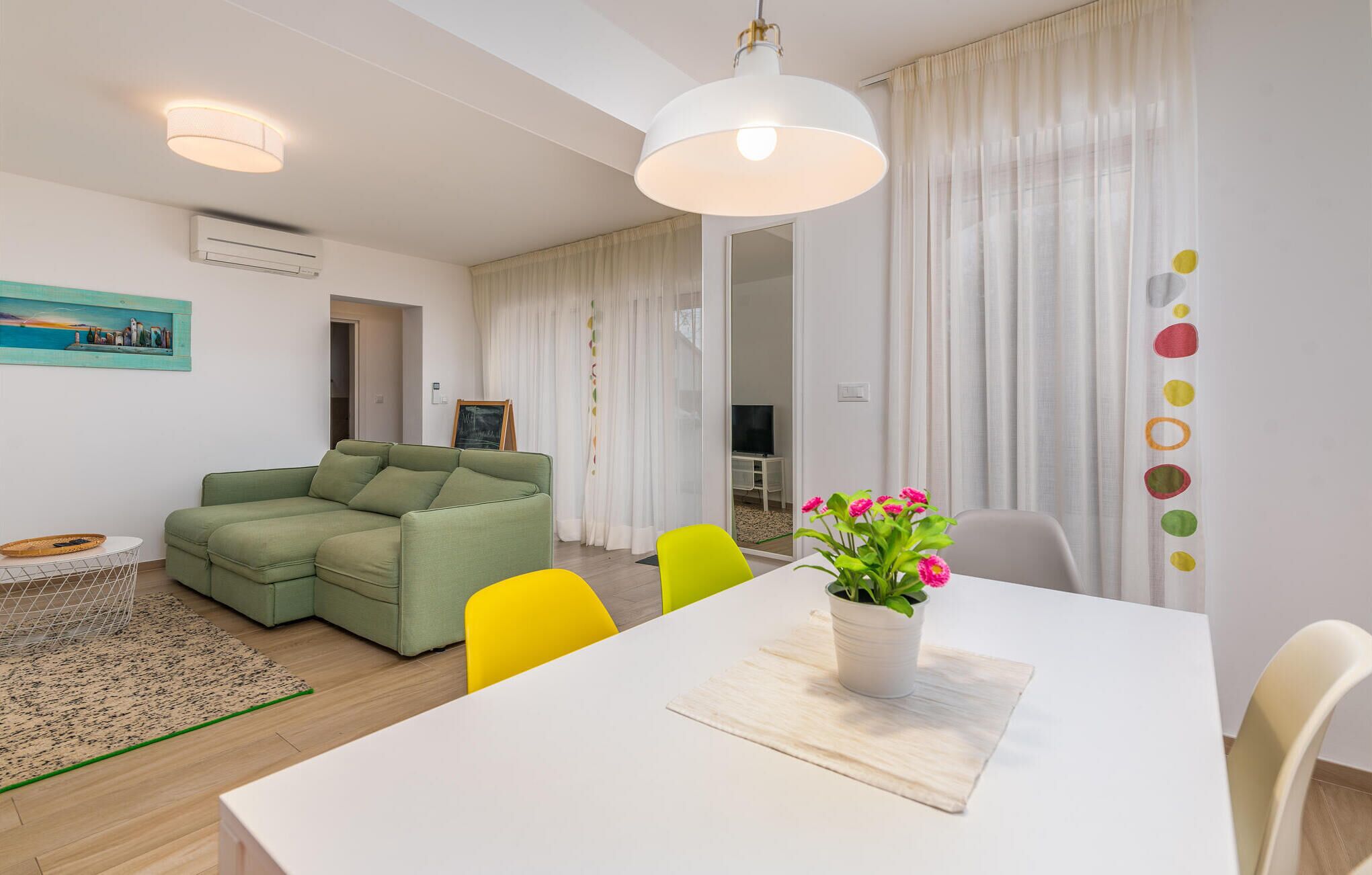Una sala de estar moderna con un sofá verde, sillas amarillas, una mesa de comedor blanca y una planta en maceta.