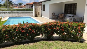 A swimming pool with a stone wall, a covered patio with chairs, and a hedge with red flowers. - Porto de Galinhas House in Cond. by the sea Esmeralda do Mar (Ipojuca)