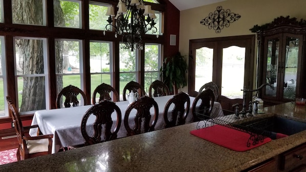 A dining area with a table set for six, surrounded by wooden chairs, a chandelier, and a view of a garden through large windows.