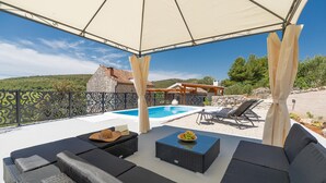 Un espace extérieur aménagé, abrité et doté d’une piscine, de chaises longues et d’une vue sur un bâtiment en pierre et des collines.