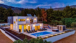 A modern two-story house with a swimming pool, outdoor seating, and a view of the surrounding forest and mountains.