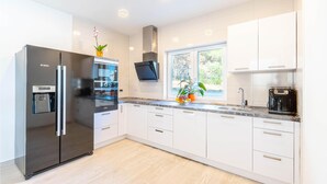 A modern kitchen with a stainless steel refrigerator, a built-in oven, a coffee machine, and a window with a view of greenery.