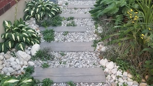 A narrow pathway flanked by brick walls and lush greenery, leading to a garden area.