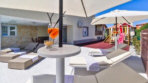 Un espace de détente extérieur baigné de soleil, avec des fauteuils en osier, une petite table ronde sur laquelle se trouve une boisson, et, au fond, une structure de jeux pour enfants.