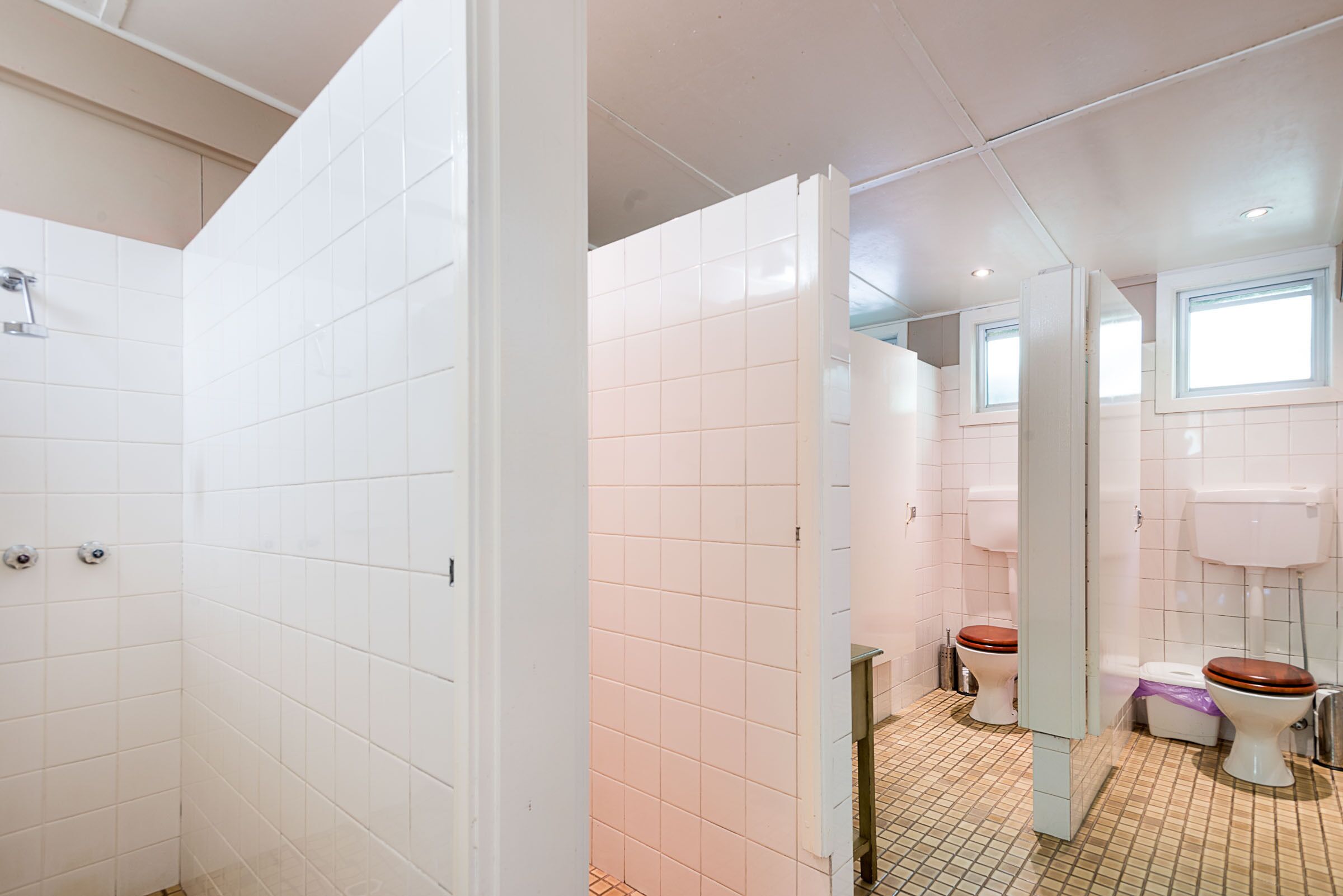 A bathroom with white tiled walls, a tiled floor, a shower area, two toilets, and a window providing natural light.