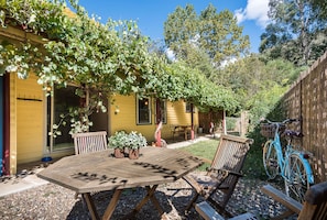 A wooden table and chairs in a garden area with a yellow building, a bicycle, and a wooden fence.