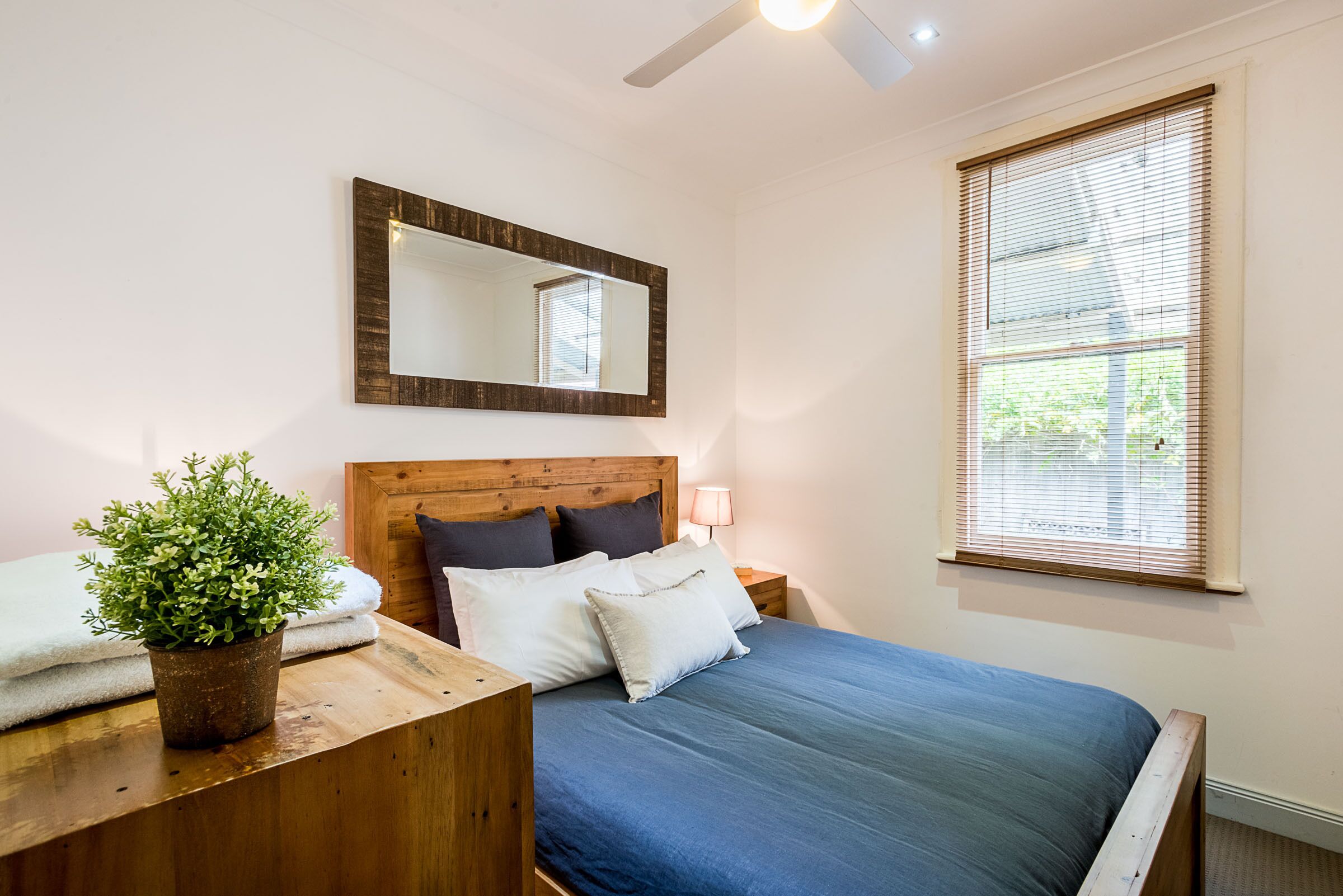 A bedroom with a wooden headboard, a large mirror, a bedside table with a plant, a blue bedspread, and a window with blinds.