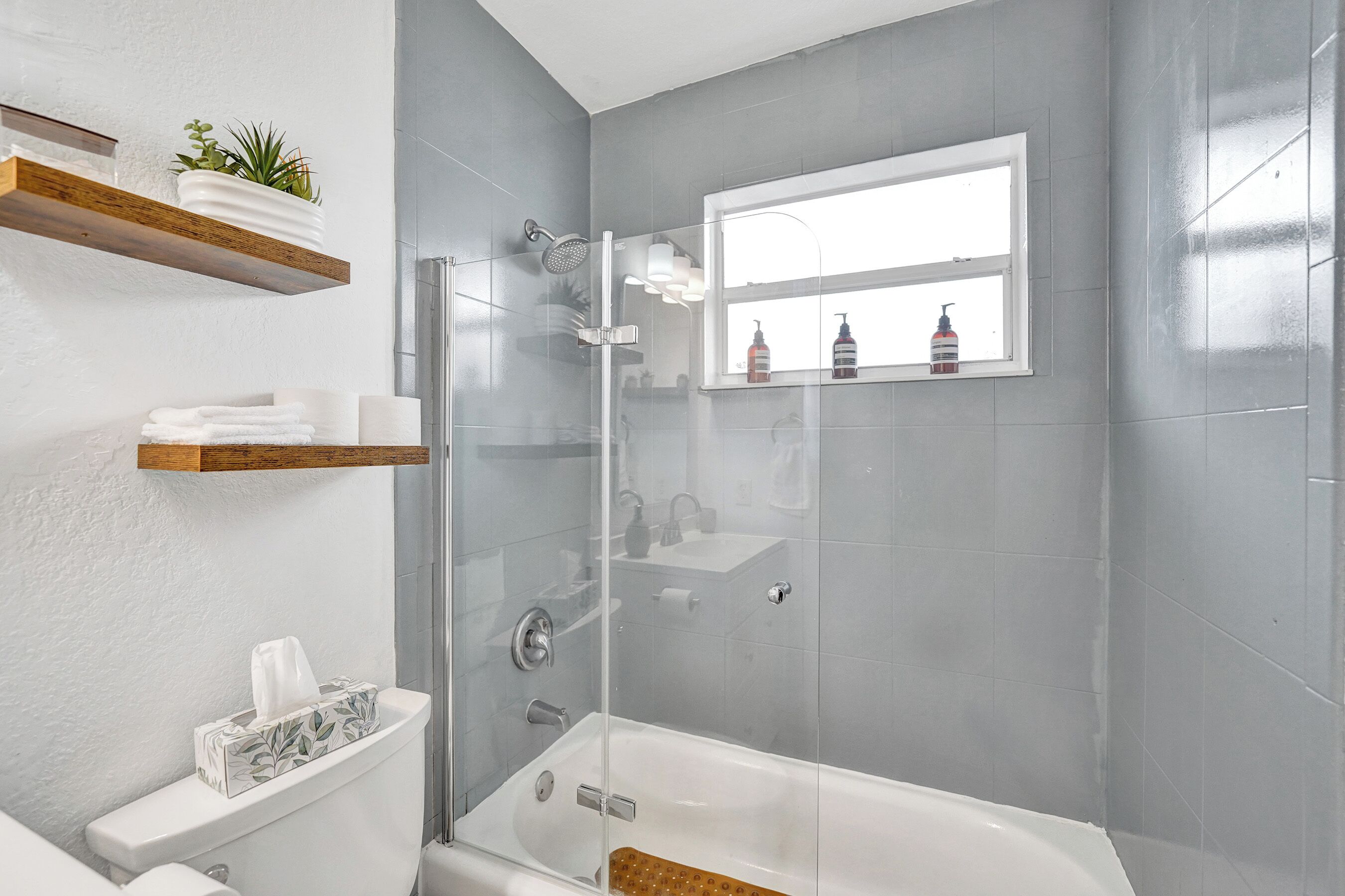 A modern bathroom with a glass-enclosed shower, a white bathtub, and a window above the sink.
