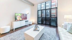 A modern living room with a large TV, white furniture, and a view of a building through a glass door. - Santana Row Properties #7 - Silicon Valley Getaway (San Jose)