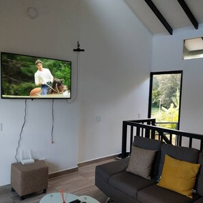 Een moderne woonkamer met een wandtelevisie, een grijze bank met kussens, een klein bijzettafeltje en een raam met uitzicht op groen.