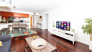 Een moderne woonkamer met een flatscreen televisie, houten vloeren, een eethoek met een tafel en stoelen, en een keuken met roestvrijstalen apparatuur.