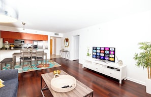 Ein modernes Wohnzimmer mit Flachbildfernseher, Holzboden, einer Essecke mit Tisch und Stühlen sowie einer Küche mit Edelstahlgeräten.
