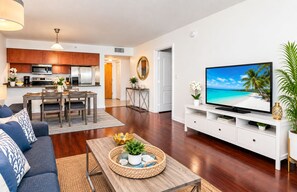 A modern living room with a flat-screen TV, a sofa, a coffee table with decorative items, and a kitchen in the background.