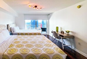 Ein Hotelzimmer mit zwei Betten, einem Schreibtisch mit Stuhl, einem kleinen Tisch mit einer Vase und Blick auf die Stadt durch große Fenster.
