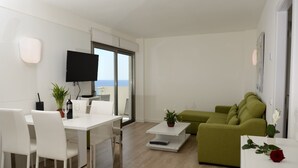 A modern living room with a white dining table, white chairs, a green sofa, and a flat-screen TV mounted on the wall.