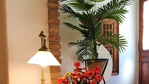 Una mesa con cubierta de vidrio, una lámpara de latón, un jarrón con flores rojas y una planta en maceta en un rincón de una habitación con pared de ladrillo y puerta de madera.