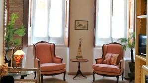 Una sala de estar acogedora con dos sillones, una mesita y un sofá. Hay dos ventanas con cortinas, una chimenea y un televisor montado en la pared.