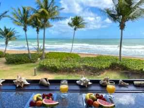 Blick auf einen tropischen Strand mit Palmen, Wellen und einem Tisch mit Früchten und Getränken.