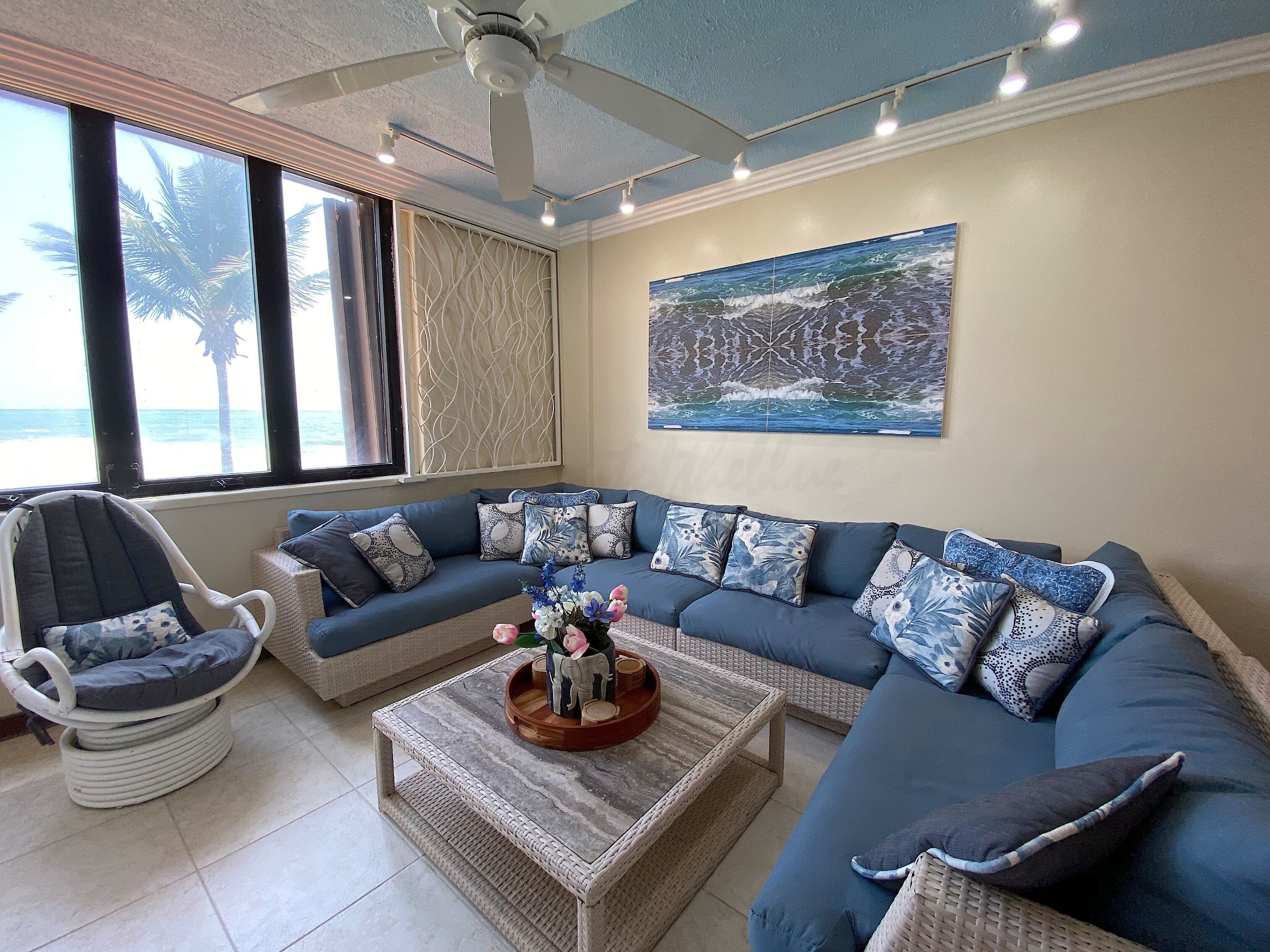 A living room with a blue sofa, a wicker chair, a coffee table with a decorative centerpiece, and a large ocean-themed painting on the wall.
