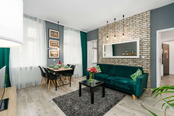 A modern living room with a green sofa, a dining table with chairs, a TV mounted on a brick wall, and a small table with a vase of flowers.