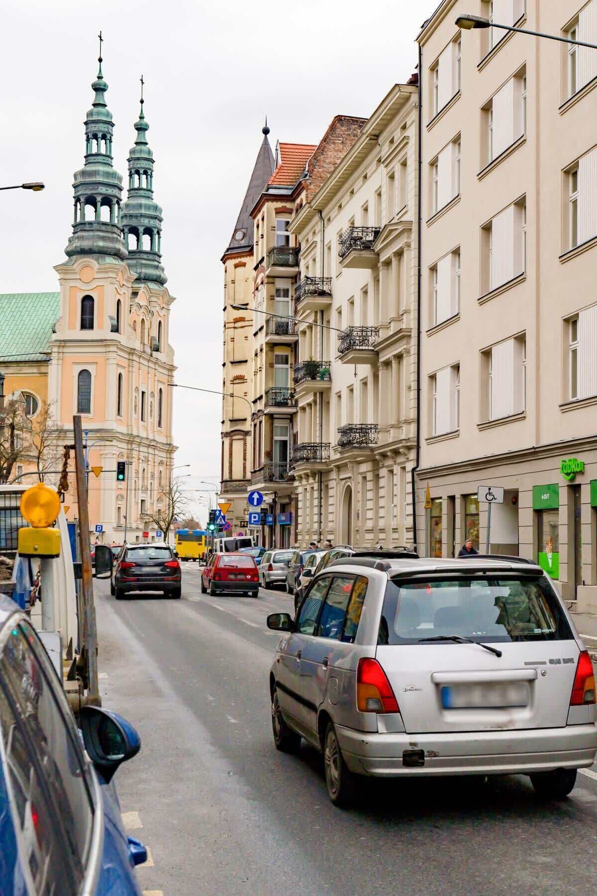 Una calle con coches, edificios y una iglesia con dos torres verdes.