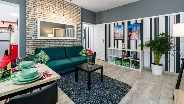 Een moderne woonkamer met een geaccentueerde bakstenen wand, een groene bank, een eethoek met een tafel voor vier personen, en een boekenplank vol boeken en decoratieve objecten.
