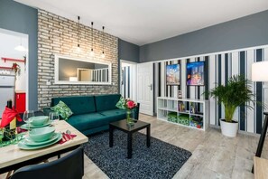 Een moderne woonkamer met een geaccentueerde bakstenen wand, een groene bank, een eethoek met een tafel voor vier personen, en een boekenplank vol boeken en decoratieve objecten.