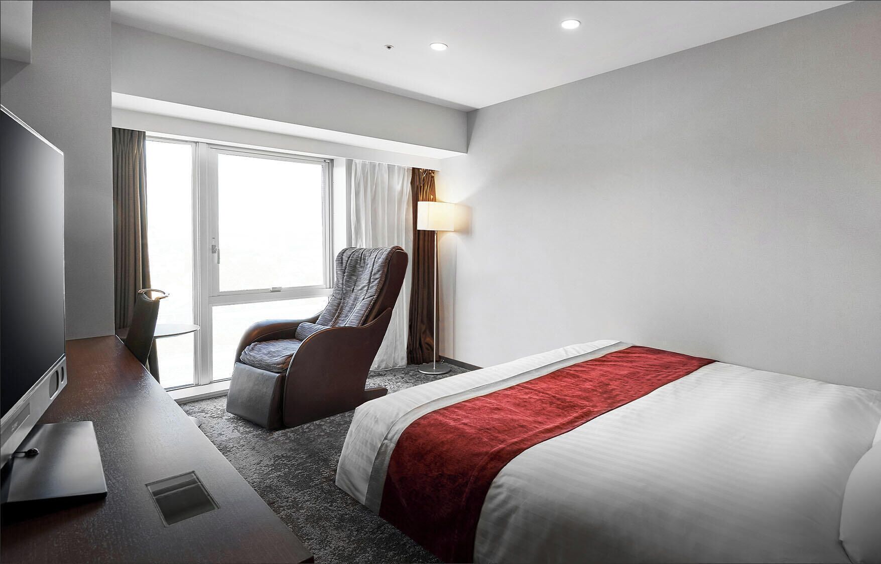 A hotel room with a bed, a desk, a chair, a lamp, and a window with curtains.