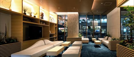 A modern hotel lobby with a large TV, comfortable seating areas, and a view of the city through large windows.