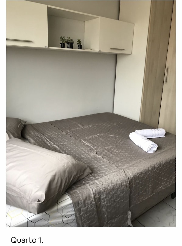 A bedroom with a bed, pillows, a folded towel, a wardrobe, and a shelf with small plants.