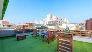 A rooftop terrace with wooden furniture and a green artificial turf, overlooking a cityscape with modern buildings. - SeeU House (Seoul)