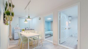 A modern kitchen with a microwave, sink, and hanging plants. There is a dining area with a white table and chairs, and a bedroom with a bed and curtains. - SeeU House (Seoul)