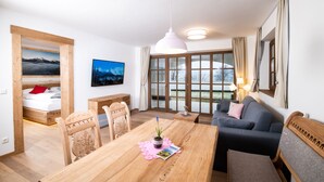 Un salon moderne avec une table à manger en bois, un canapé et un lit agrémenté d’un coussin rouge. Deux tableaux ornent le mur, et une grande fenêtre est habillée de rideaux.