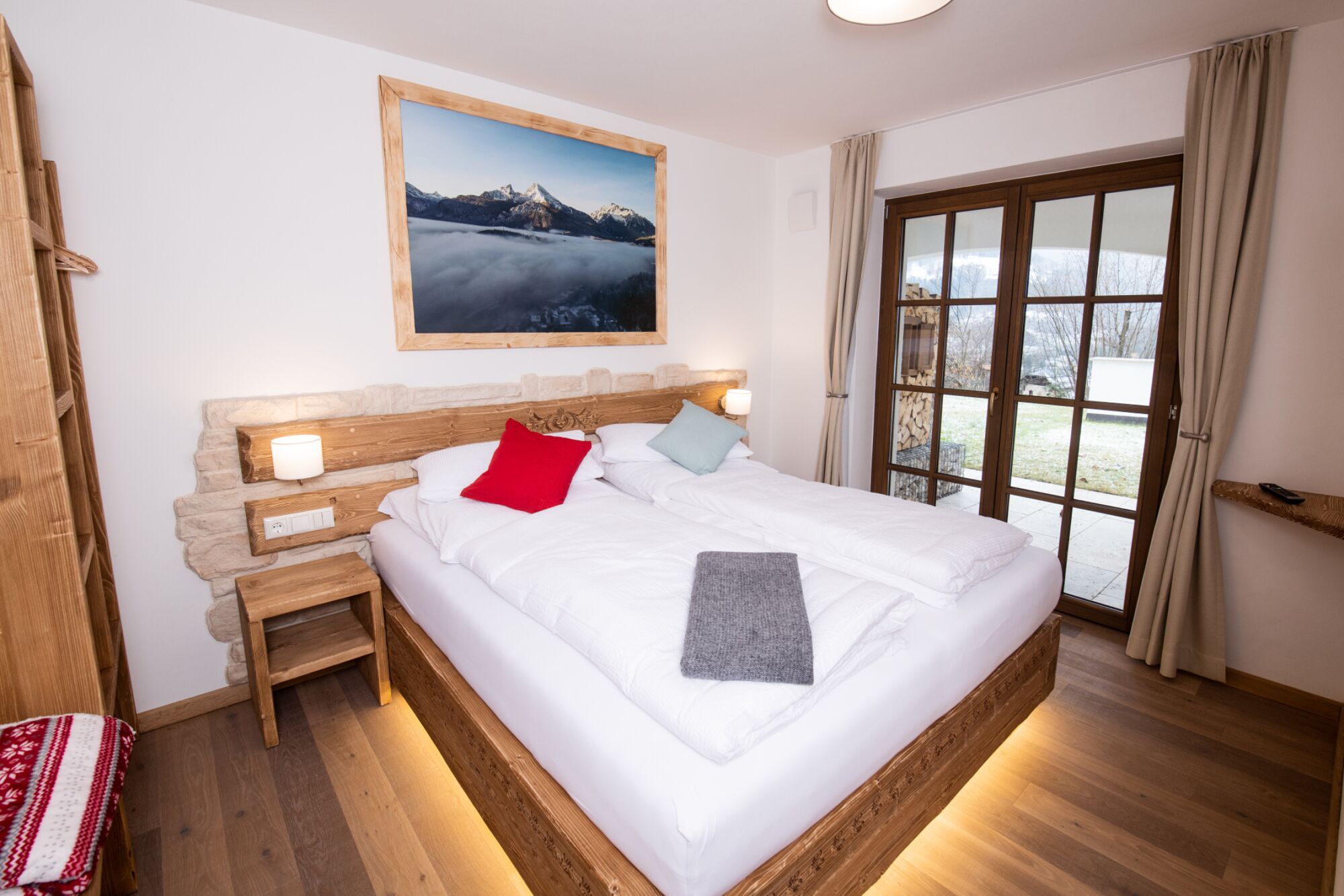 Ein Schlafzimmer mit einem hölzernen Kopfende, einem großen Fenster mit Vorhängen, einem Nachttisch mit Lampe und einem Gemälde über dem Bett.