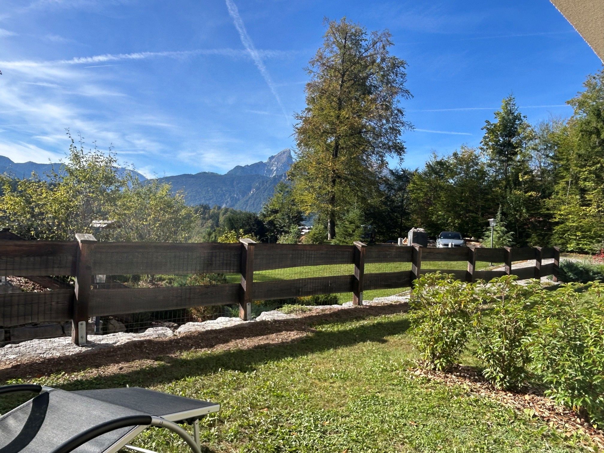 Ein von einer Hecke umgebener Außenbereich mit einer Bank und einem Tisch, im Hintergrund Bäume und Berge.