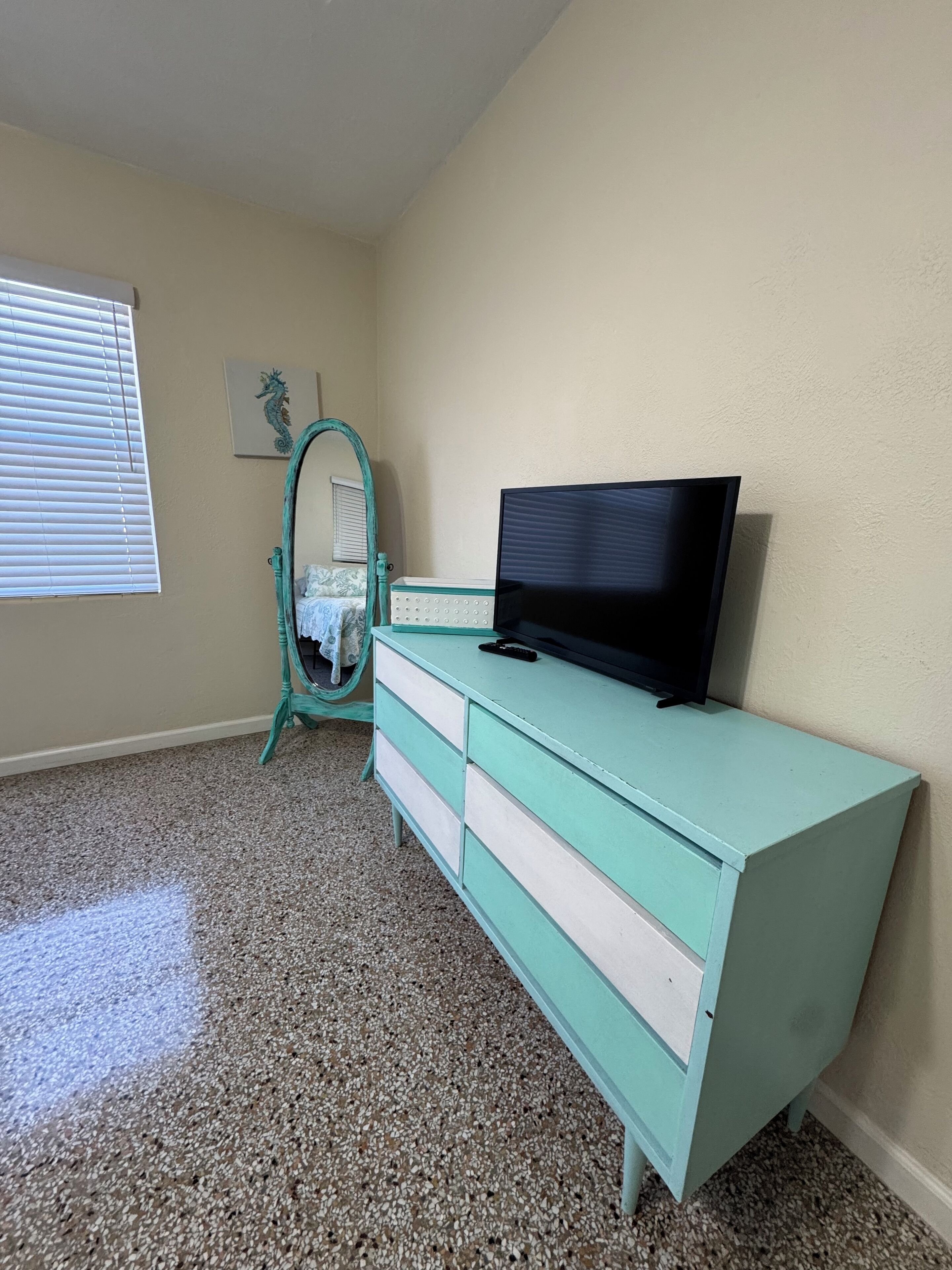 A room with a teal dresser, a television on top, a mirror, and a window with blinds.