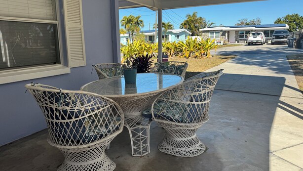 A covered patio with a round table and wicker chairs, overlooking a street with parked cars and houses.