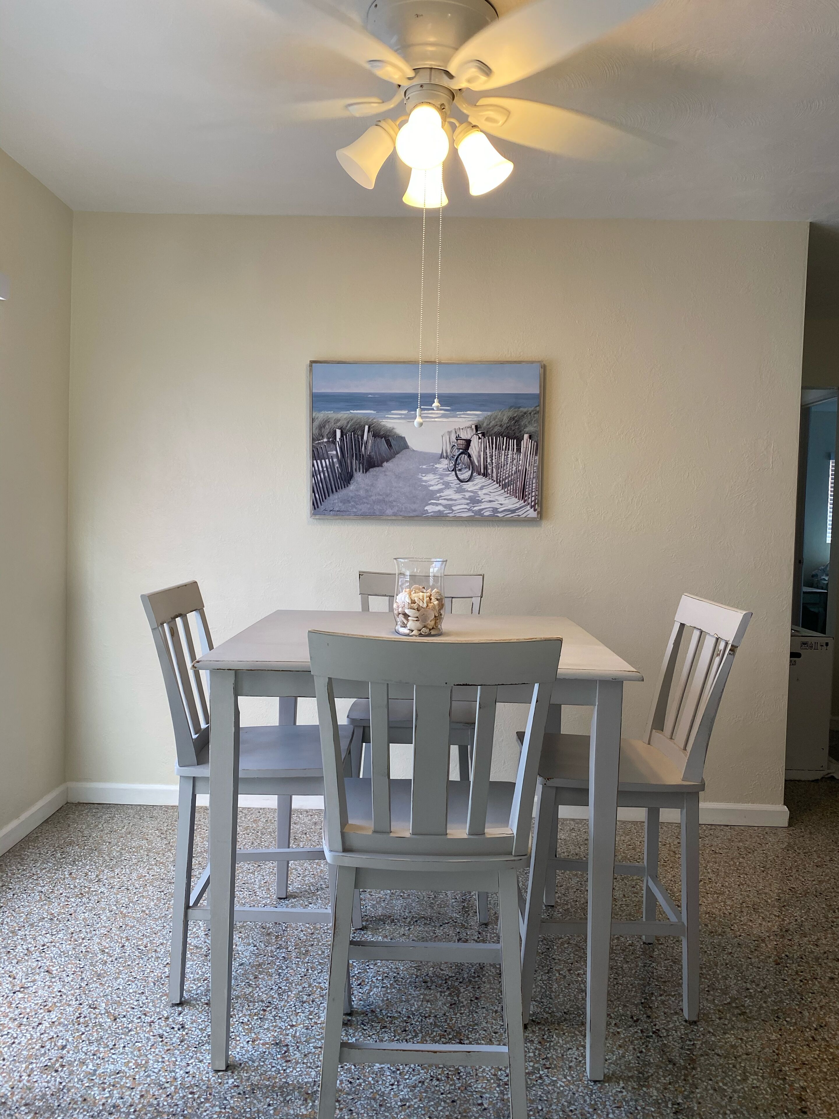 A dining area with a table and four chairs, a ceiling fan, and a beach-themed painting on the wall.