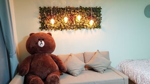 Ein Zimmer mit einem großen braunen Teddy auf einem hellen Sofa, einer wandmontierten Leuchte mit Blättern und einem Bett mit einem gemusterten Bettbezug.