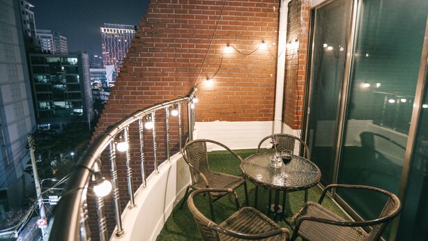 Ein Balkon mit Korbstühlen und einem kleinen Tisch, mit Blick auf eine nächtliche Stadtlandschaft.