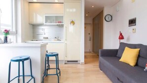 A modern kitchen with white cabinets, a microwave, and a sink. There are two blue stools and a gray sofa with a yellow pillow. A clock is on the wall. - Sweet House2 (Seoul)