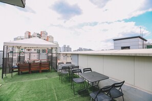 A rooftop terrace with a wooden bench, tables, and chairs, surrounded by a white wall and a view of buildings in the background. - Lovely House (Seoul)