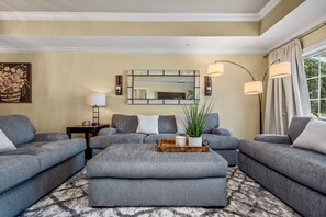 Un salon avec des canapés gris, un pouf central, un miroir et une lampe sur pied.