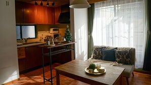 A cozy living room with a wooden table, a sofa with patterned cushions, a small Christmas tree, and a kitchen area with wooden cabinets and a microwave. - Ikken Rakutei (Tokyo)