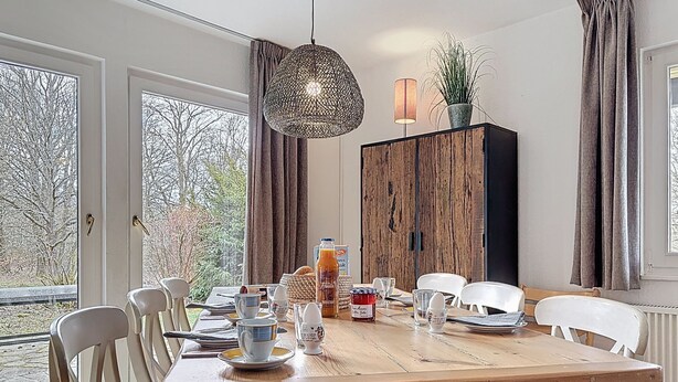 Un comedor con mesa de madera, sillas blancas, lámpara colgante y vistas amplias por ventanas grandes.